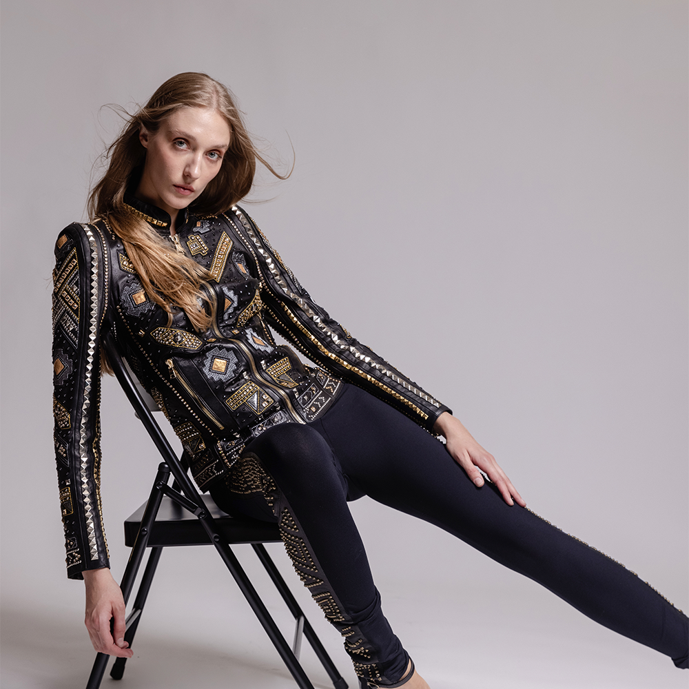 Woman wearing an ornate black and gold jacket sitting on a chair against a gray background